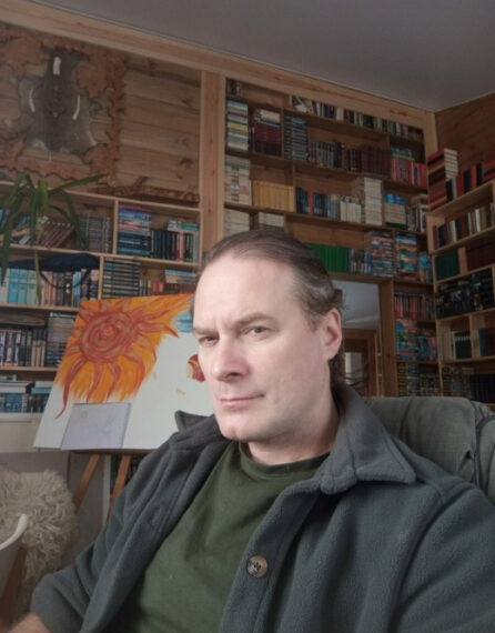 Мужчина в комнате с книжными полками, рядом картина с изображением подсолнуха.