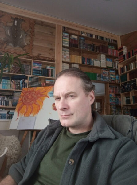 Мужчина в комнате с книжными полками, рядом картина с изображением подсолнуха.