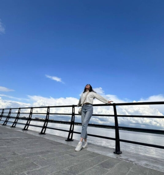 Девушка на набережной в солнечный день, облокотившись на перила, небо и облака на фоне.