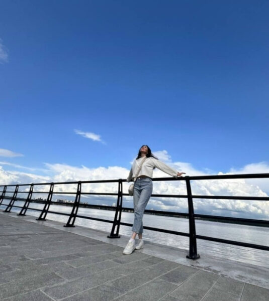 Девушка на набережной в солнечный день, облокотившись на перила, небо и облака на фоне.