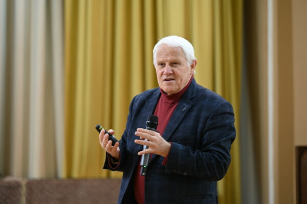 «Через 20–30 лет мы действительно не узнаем наш мир 5 Мужчина с микрофоном выступает на фоне жёлтых штор.