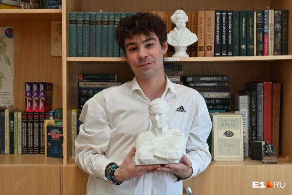 Человек в белой рубашке держит бюст и стоит у книжного шкафа.