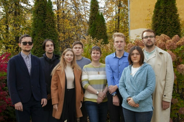 Группа людей на улице осенью, позируют на фоне зелени и жёлтой стены.