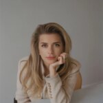 Блондинка в бежевом пиджаке за столом, светлый интерьер.