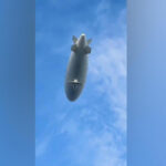 Дирижабль в голубом небе, вид снизу, летит высоко над облаками.