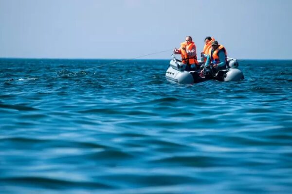 Двое рыбаков в лодке на синем море в оранжевых спасательных жилетах ловят рыбу.