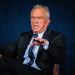 Мужчина в костюме сидит на стуле, жестом выделяя мысль, с серьезным выражением лица.