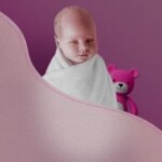 Младенец в белом одеяле, фиолетовый фон с розовым плюшевым медведем.