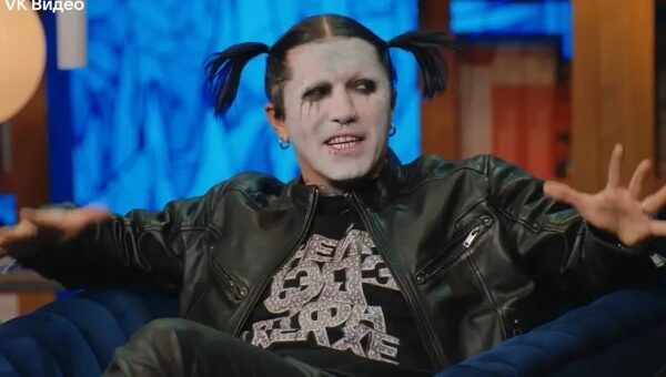 Человек с ярким макияжем и двумя хвостиками в кожаной куртке на синем фоне.