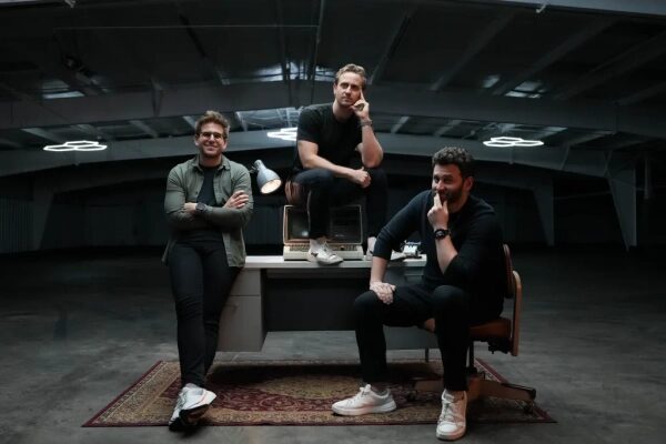 Трое мужчин позируют у стола в стильном помещении с индустриальным дизайном.