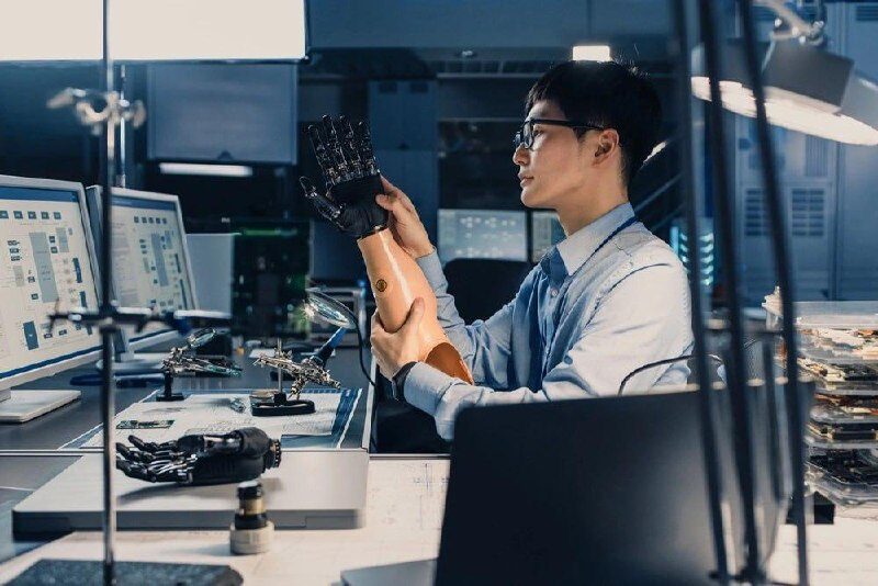 Инженер исследует протез в лаборатории, окруженной компьютерами и робототехникой.