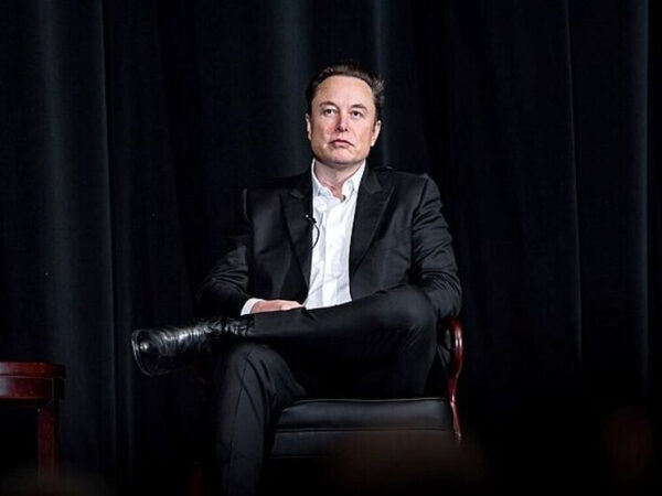 Мужчина в черном костюме сидит на стуле на сцене с черным фоном.