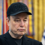 Человек в черной кепке с надписью "DOGE" на фоне цветных занавесок и флага США.