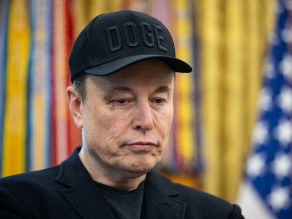 Лунные фантазии Маска: что стоит за проектом первого Лунограда 11 Человек в черной кепке с надписью "DOGE" на фоне цветных занавесок и флага США.