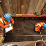 Археологи в защитных касках и жилетах работают на раскопках в траншее.