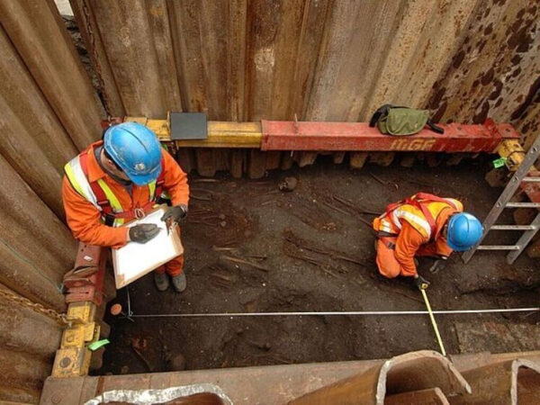 Археологи в защитных касках и жилетах работают на раскопках в траншее.