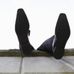 Мужчина в костюме лежит на стене, видны только его ноги в черных туфлях на фоне неба.