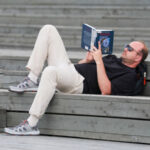 Мужчина лежит на деревянных ступенях и читает книгу на свежем воздухе.