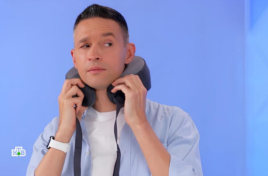 Мужчина в голубой рубашке держит наушники на фоне синей стены.