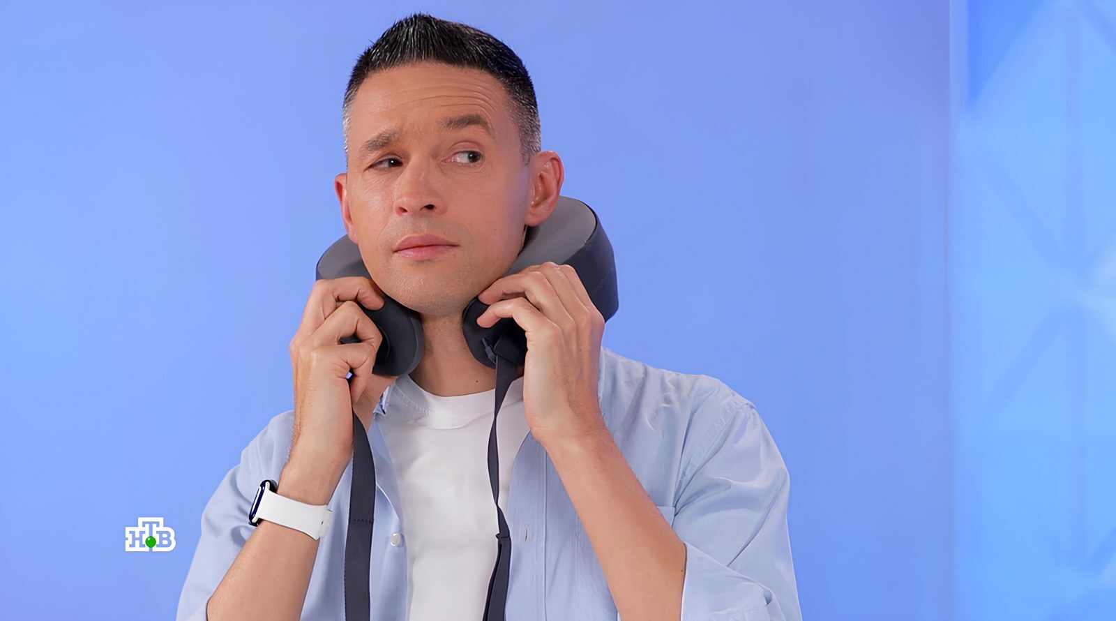Мужчина в голубой рубашке держит наушники на фоне синей стены.