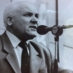 Мужчина в костюме на черно-белой фотографии говорит в микрофон на фоне стены.