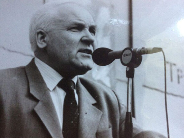 Мужчина в костюме на черно-белой фотографии говорит в микрофон на фоне стены.