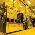 Чистая комната полупроводникового завода с оборудованием и сотрудниками в защитной одежде.