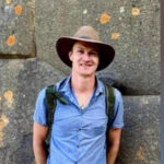 Человек в синей рубашке и шляпе с рюкзаком на фоне каменной стены.