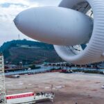 Большой дирижабль на земле с конструкциями на фоне гор и мостов.