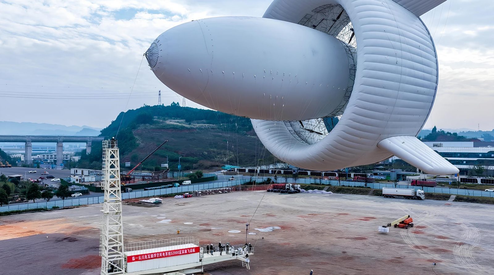 Большой дирижабль на земле с конструкциями на фоне гор и мостов.