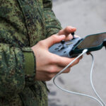 Человек в камуфляже управляет дроном с помощью пульта и телефона.