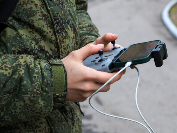 Человек в камуфляже управляет дроном с помощью пульта и телефона.