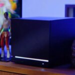 Черная квадратная колонка на тумбе дома, рядом фигурка, синий фон.