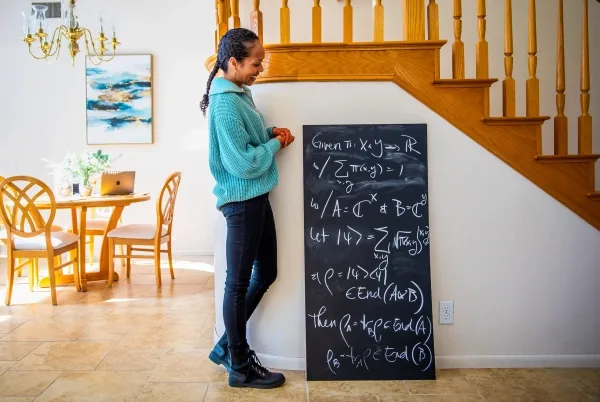 Женщина стоит рядом с доской, исписанной математическими обозначениями.