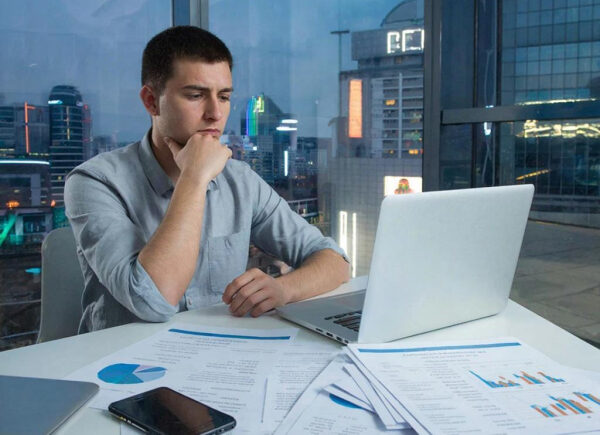 Человек работает за ноутбуком в офисе, вокруг документы и смартфон. Вид на город.