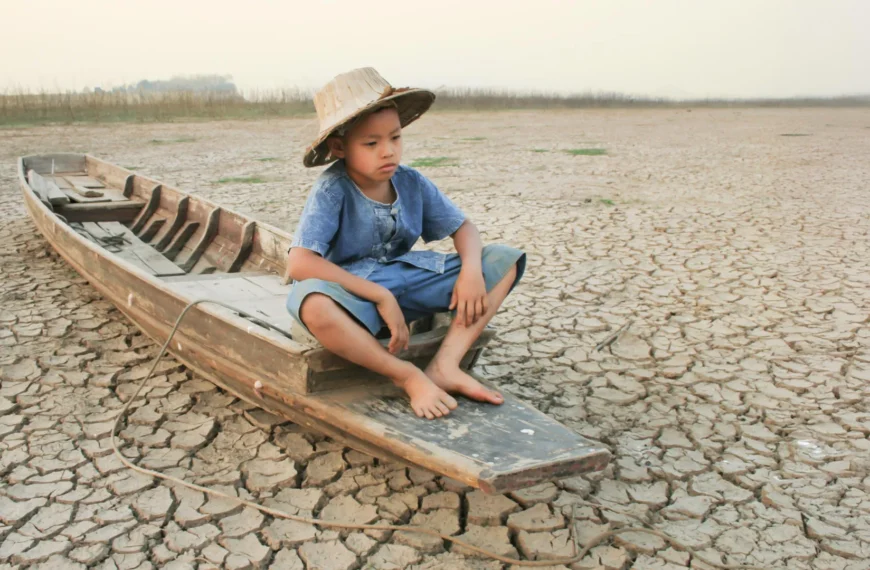 Ребёнок в шляпе сидит в лодке на высохшей земле, символизируя засуху.