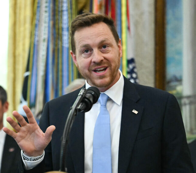 Мужчина в костюме выступает с речью у микрофона, формальная обстановка.