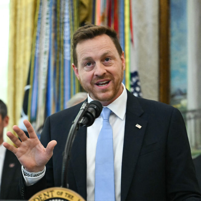 Мужчина в костюме выступает с речью у микрофона, формальная обстановка.