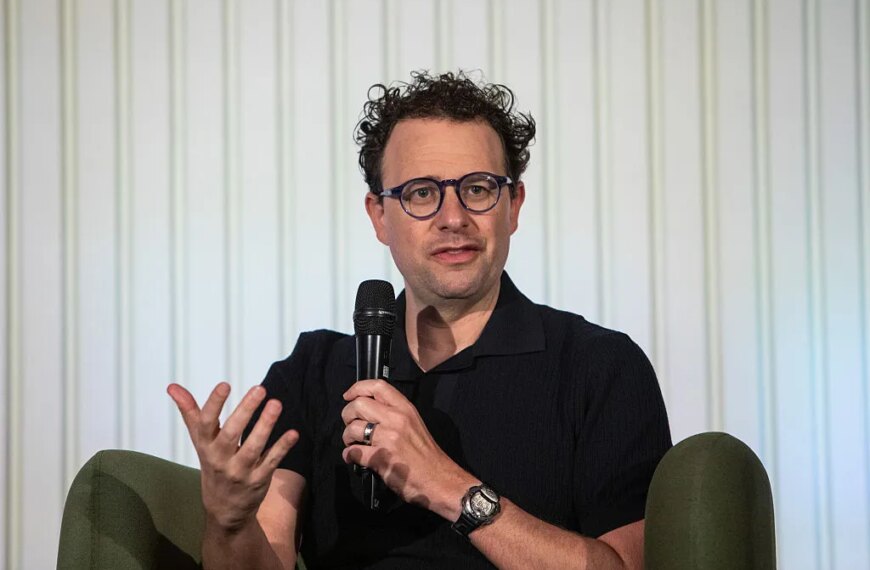 Мужчина с микрофоном в руке на конференции, делится мнением, сидя на стуле.