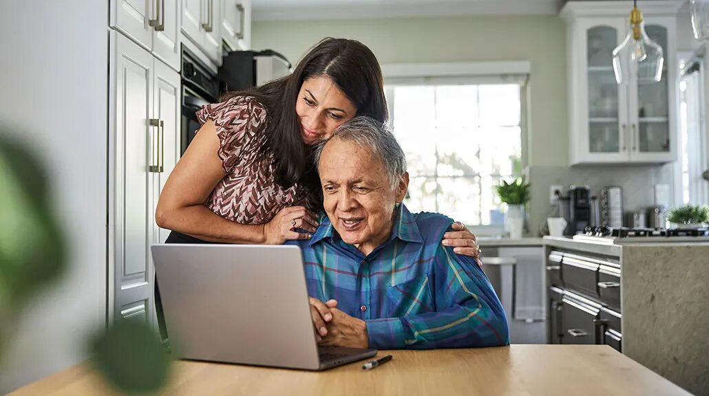 женщина средних лет склоняется над пожилым мужчиной за компьютером на солнечной кухне