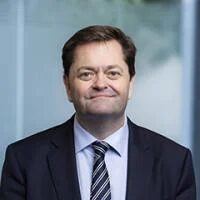 Мужчина в деловом костюме и полосатом галстуке на фоне офисной стены.