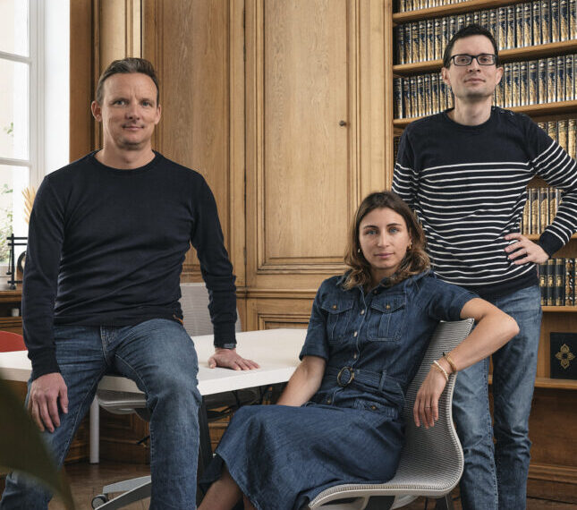 Трое людей позируют в офисе, сидят и стоят перед книжным шкафом в деловой обстановке.