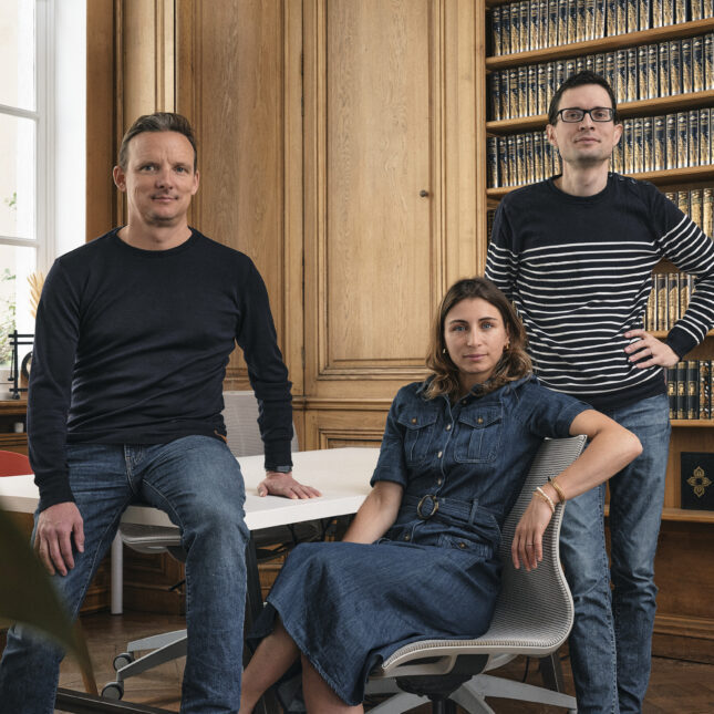 Трое людей позируют в офисе, сидят и стоят перед книжным шкафом в деловой обстановке.