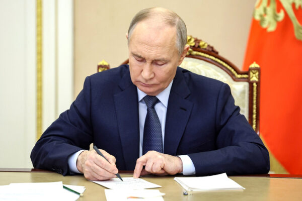 Мужчина в деловом костюме за столом подписывает документы, на заднем плане российский флаг.