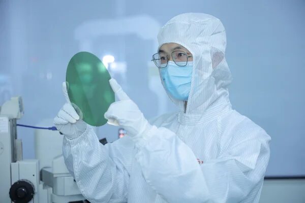 Инженер в чистой комнате исследует полупроводниковую пластину в защитном костюме.