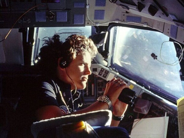 Астронавт в кабине шаттла, смотрит в иллюминатор, наушники, солнечный свет.