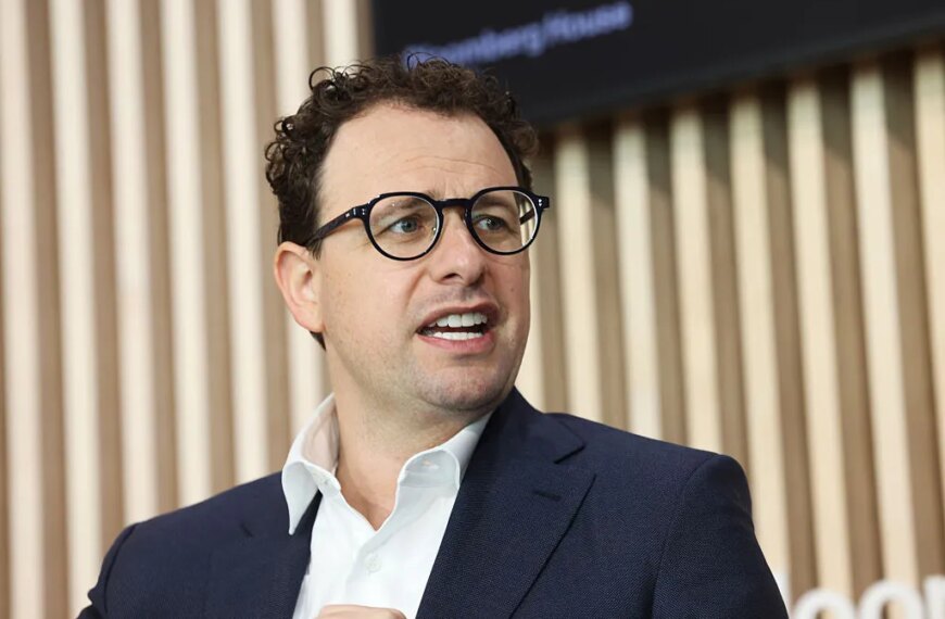 Мужчина в белой рубашке и темном пиджаке на фоне деревянных панелей.