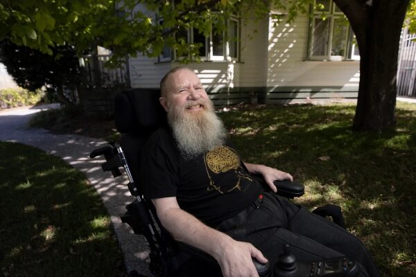 Мужчина с бородой в инвалидной коляске улыбается на улице возле дома.