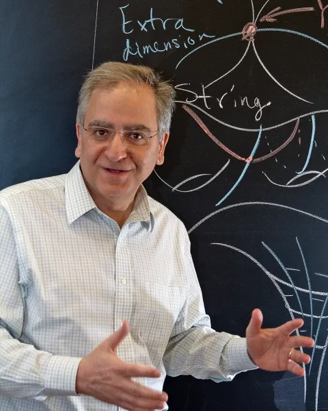 Мужчина в белой рубашке жестикулирует, как будто что-то объясняет, стоя перед доской.