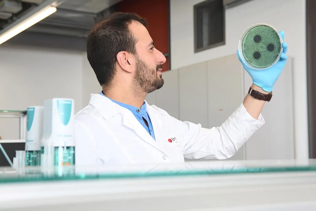 Бледный мужчина с темными волосами и бородой держит перед зрителем большую чашку Петри с тремя темными кругами